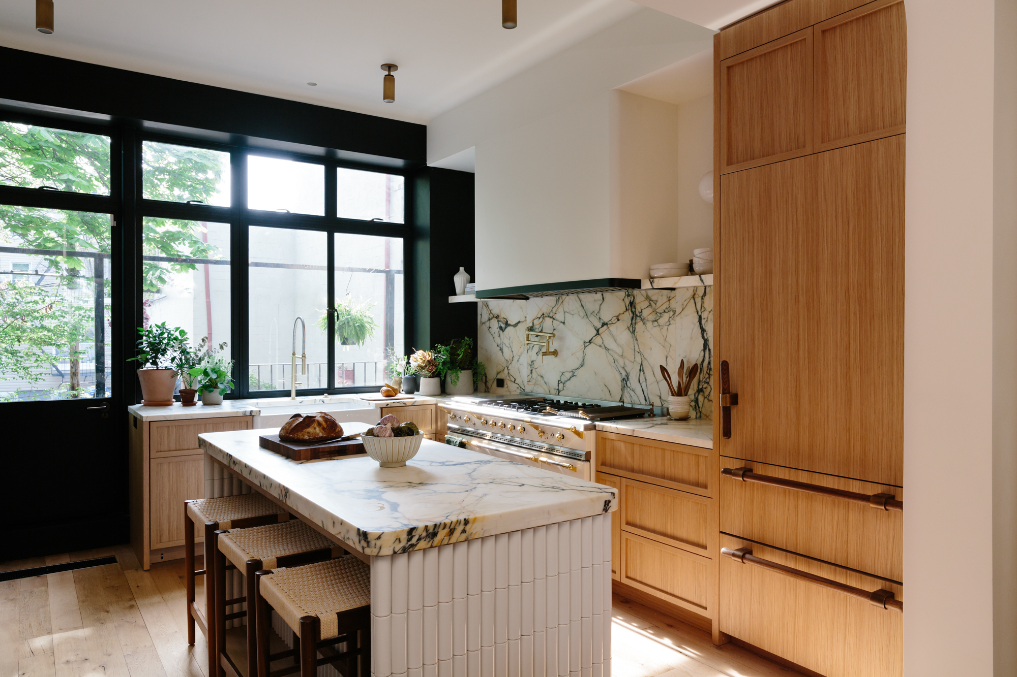 modern kitchen with timber cabinetry, marble countertops, white freestanding oven, floating marble shelves, a butlers sink, potted herbs, wall sconces, and a marble kitchen island