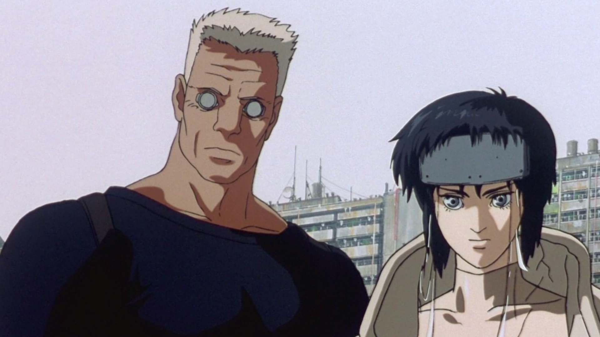 Two people, on the left is a broad-shouldered man wearing two eyepieces and a black top and on the right is someone with longer dark hair and a more defined jawline, both animated and looking back at the camera