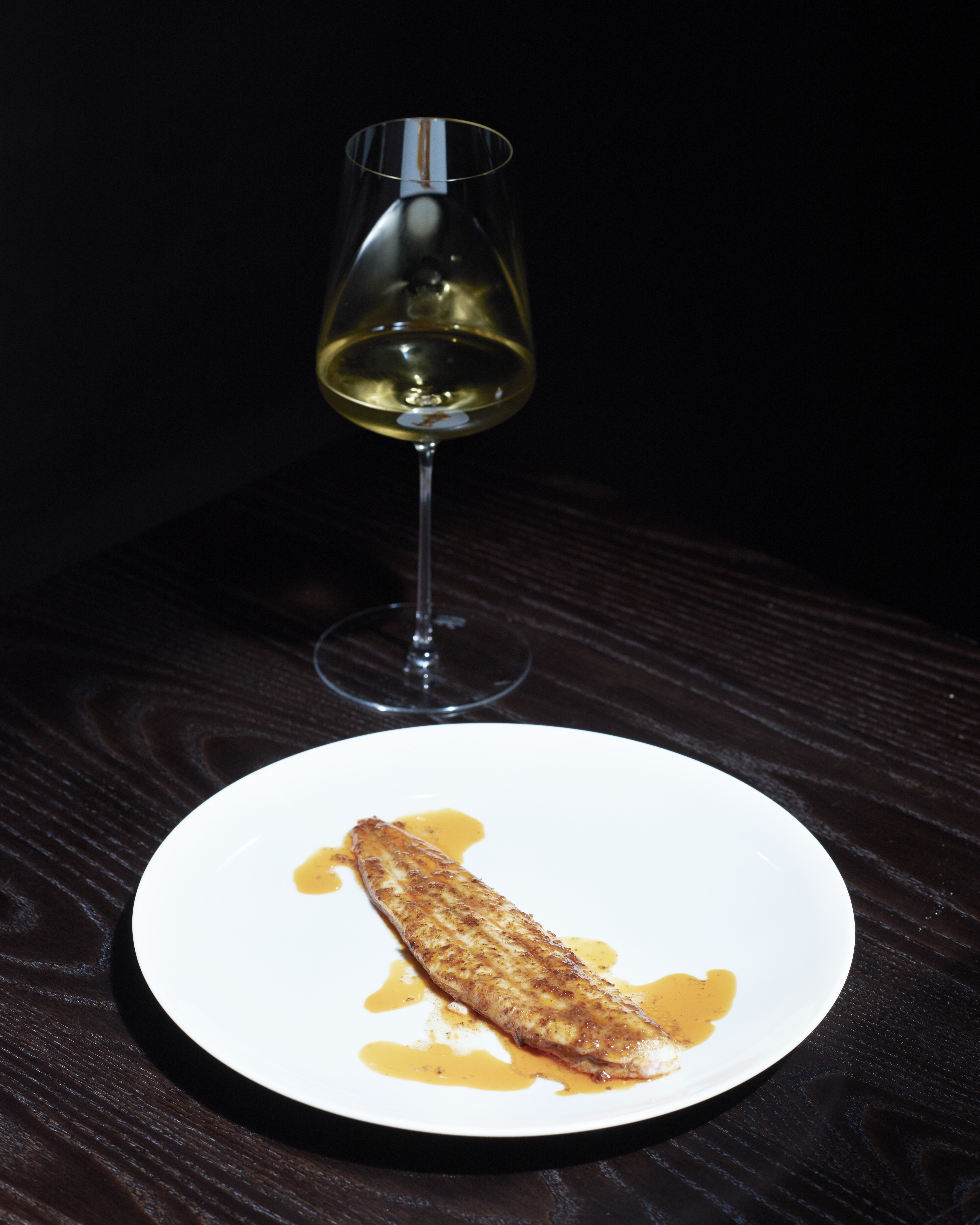 A split sole plated in a white dish on a black wooden table, next to a white wine glass.