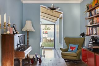 The study is a transitional space between the living room and the kitchen. Painted blue to match the kitchen, it has a piano and a bookshelf with a mid century sideboard and wingback armchair