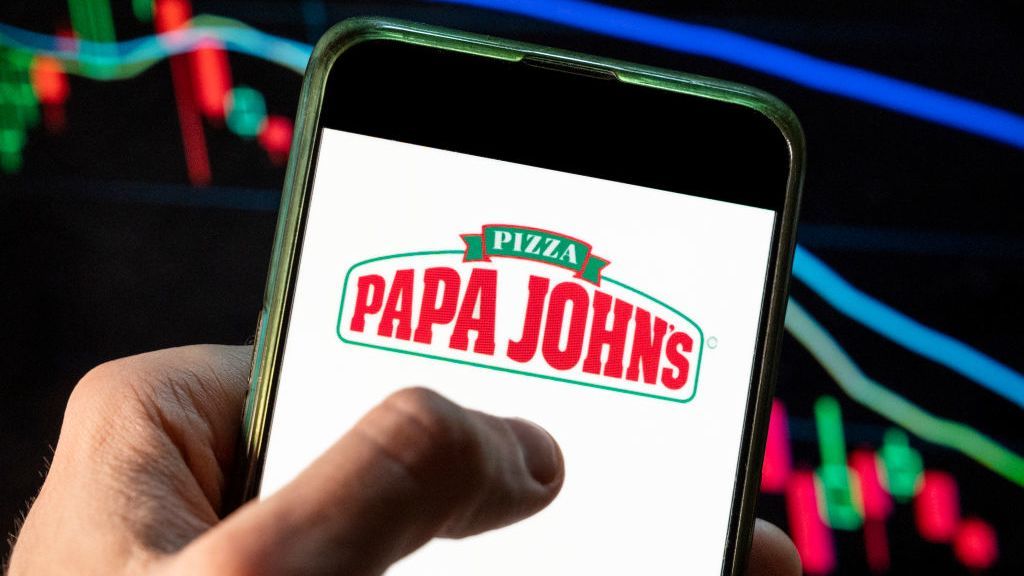 The logo of Papa John&amp;#039;s is shown on a phone held in a person&amp;#039;s hand, with a digital chart made of red, green and blue lines shown in the background