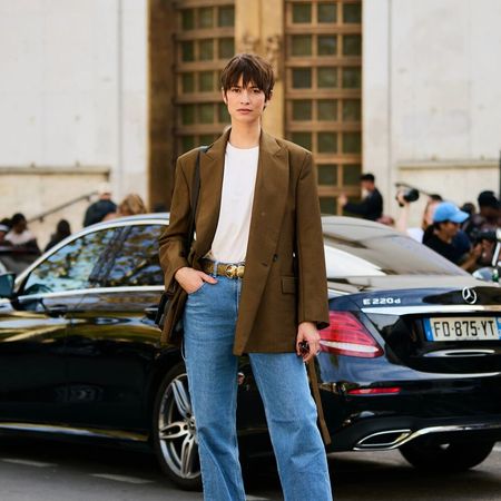 A woman wears jeans, white t-shirt, and a brown blazer while posing in the street. 