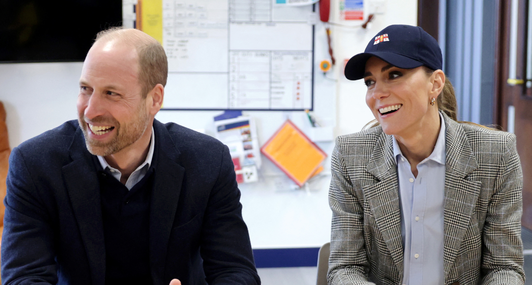 Princess Kate smiling in a baseball cap sitting next to Prince William at a table