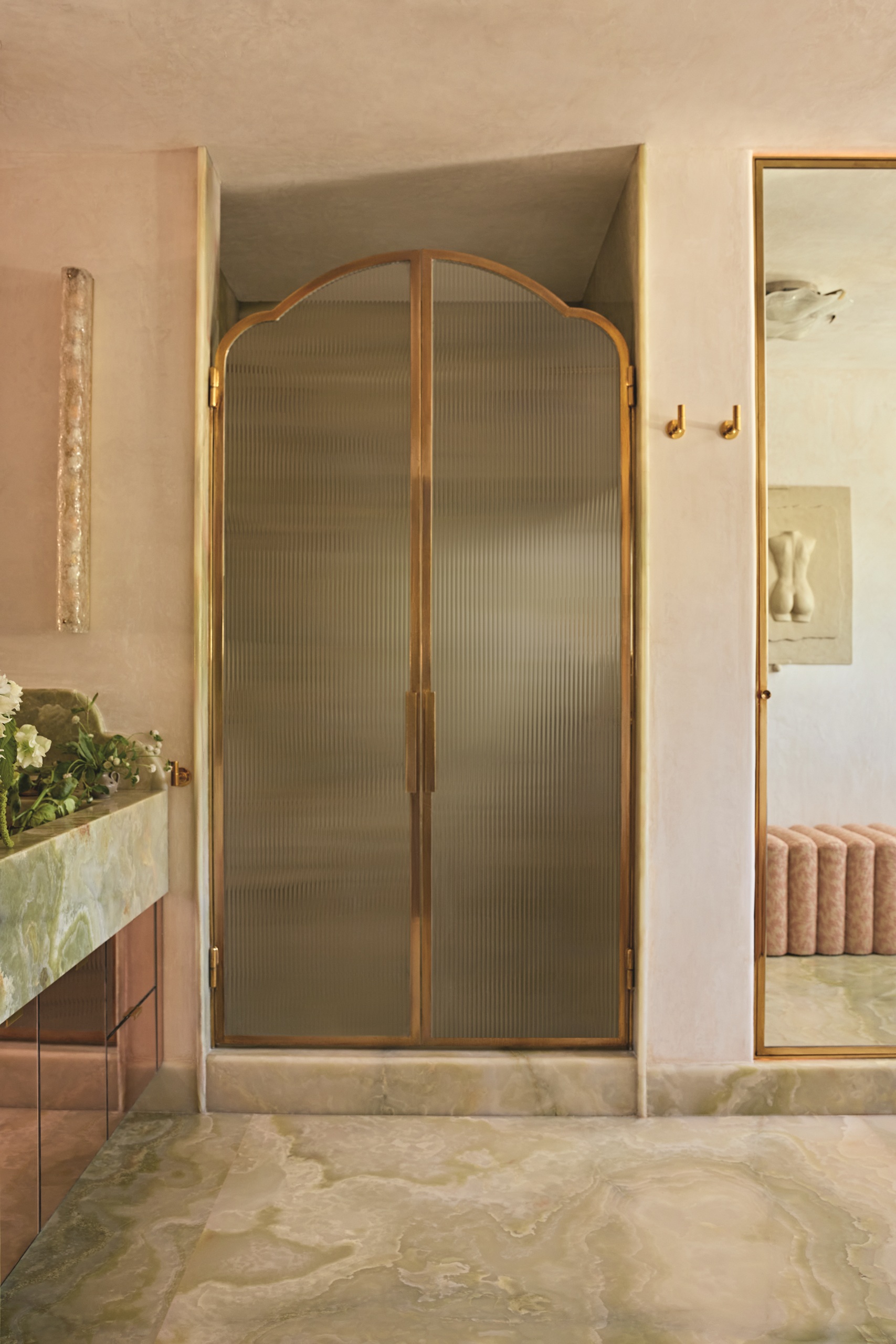 modern bathroom with marble floor, pink plaster walls, marble vanity with flowers in the sink, wall sconce, and enclosed shower with fluted glass doors with gold trim and mirror on wall