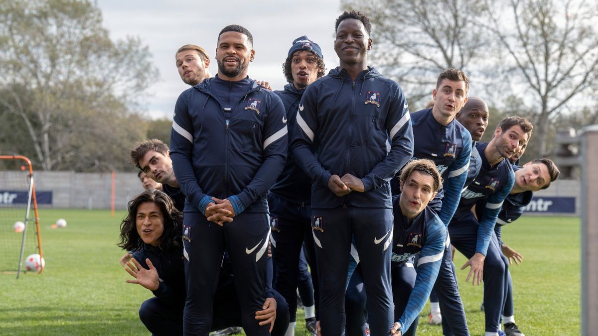 A press image of Cristo Fernández, Stephen Manas, David Elsendoorn, Kola Bokinni, Moe Hashim, Toheeb Jimoh, Phil Dunster, Billy Harris and Moe Jeudy-Lamour in &quot;Ted Lasso.&quot; performing a song from The Sound of Music on the pitch.