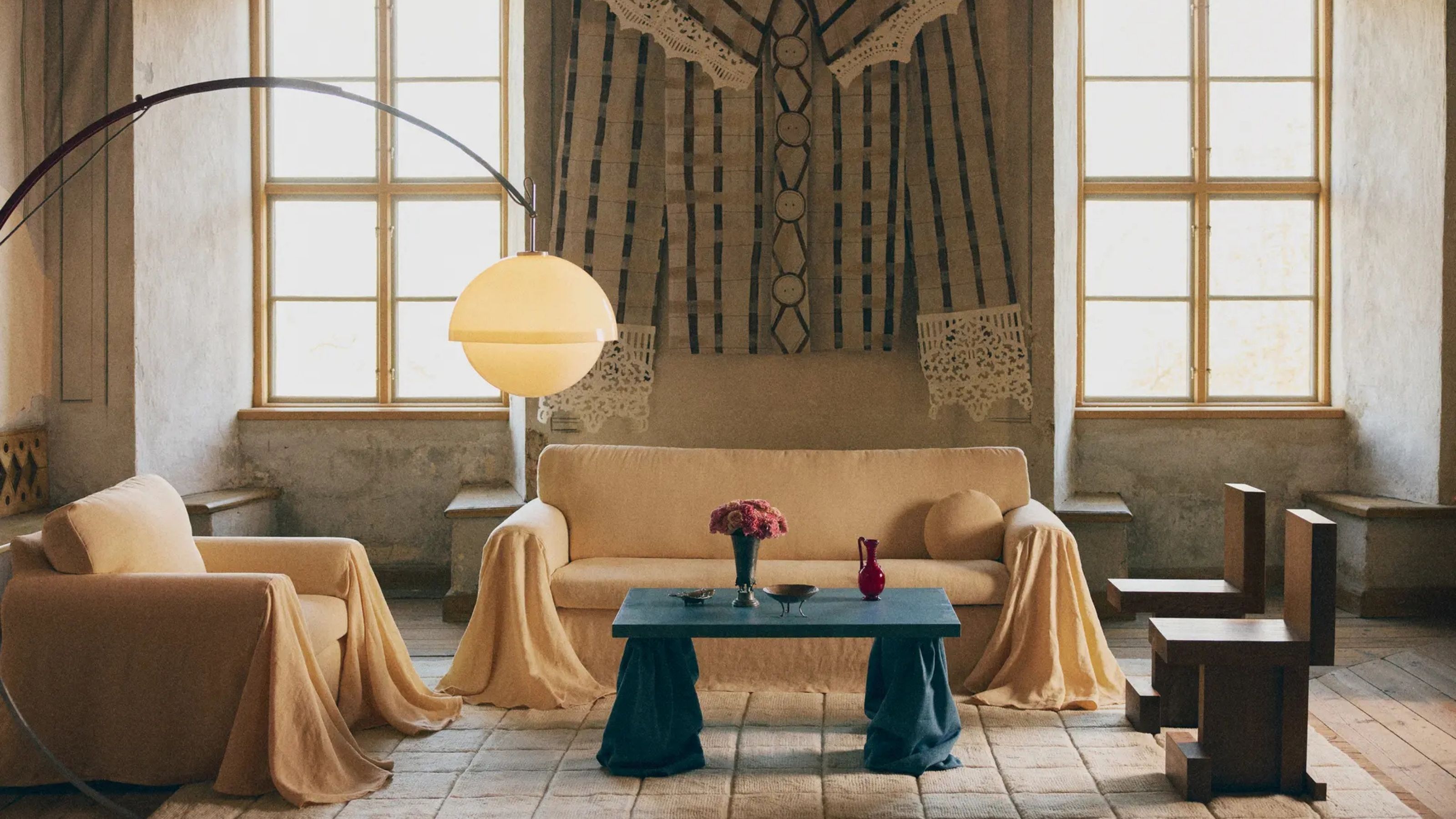 Image of a large living room with high ceilings and two large windows. There is a sofa with a pale yellow cover and draping arms, and a matching accent chair. There is a blue coffee table as well.