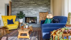 Colorful, vibrant living room with blue sofa, yellow armchair, wooden coffee table, patterned rug and plants, pictured in front of gray brick-style wall, fireplace and built-in log storage.
