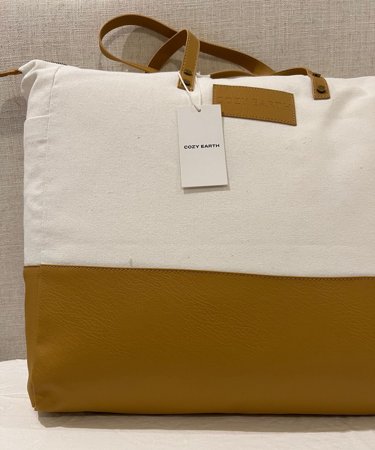 Cozy Earth Silk Comforter in a white cotton and brown leather tote bag with leather handles and a leather logo, pictured on a white bedsheet. Hanging from the left handle is a small white Cozy Earth tag. The bag is pictured against a pale, whiteish grey headboard.