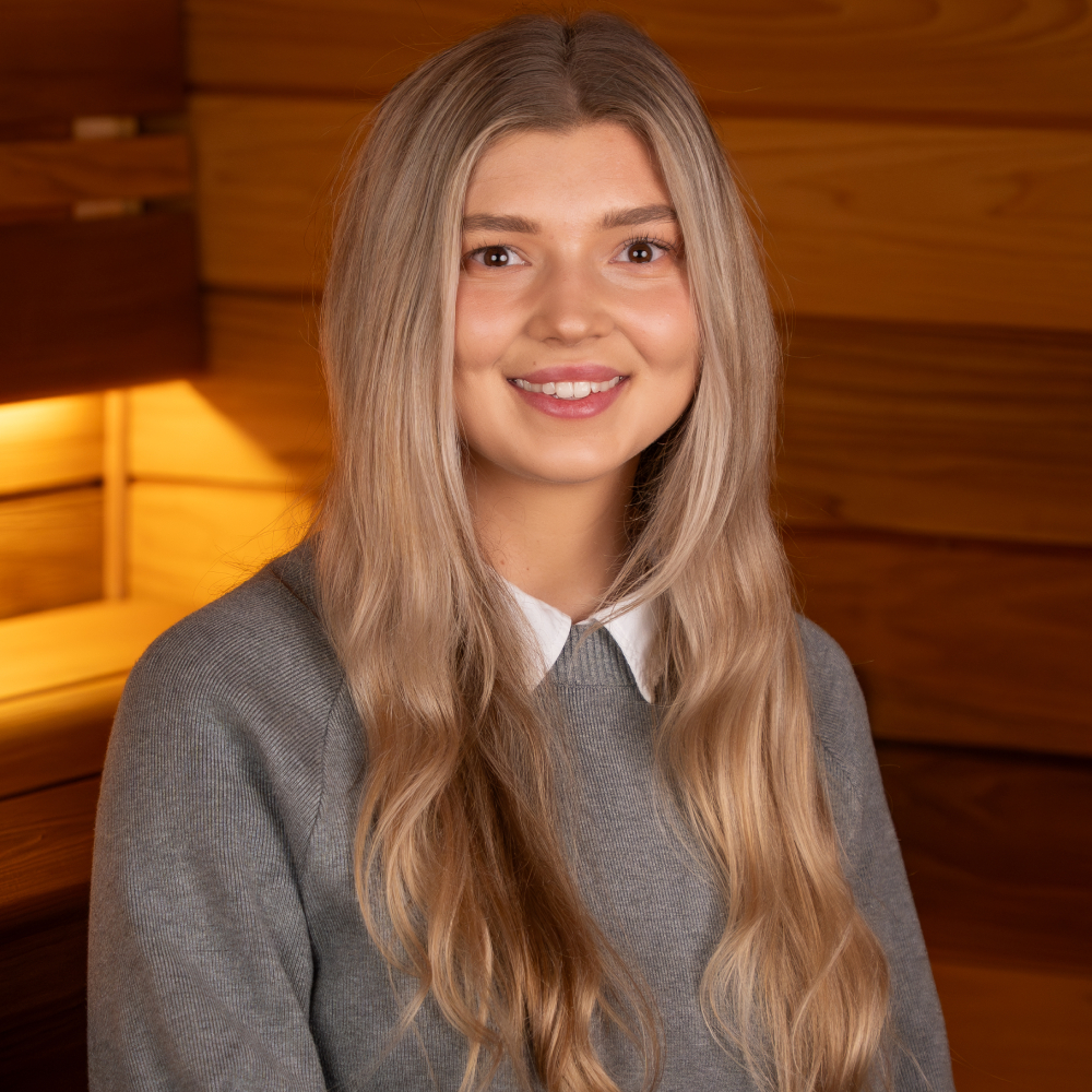 female with long blond hair wearing grey jumper and shirt sat inside sauna
