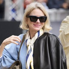 Naomi Watts in in San Sebastian, Spain wearing a leather bomber jacket, neck scarf, light blue sweater, bootcut jeans, and Adidas Samba sneakers