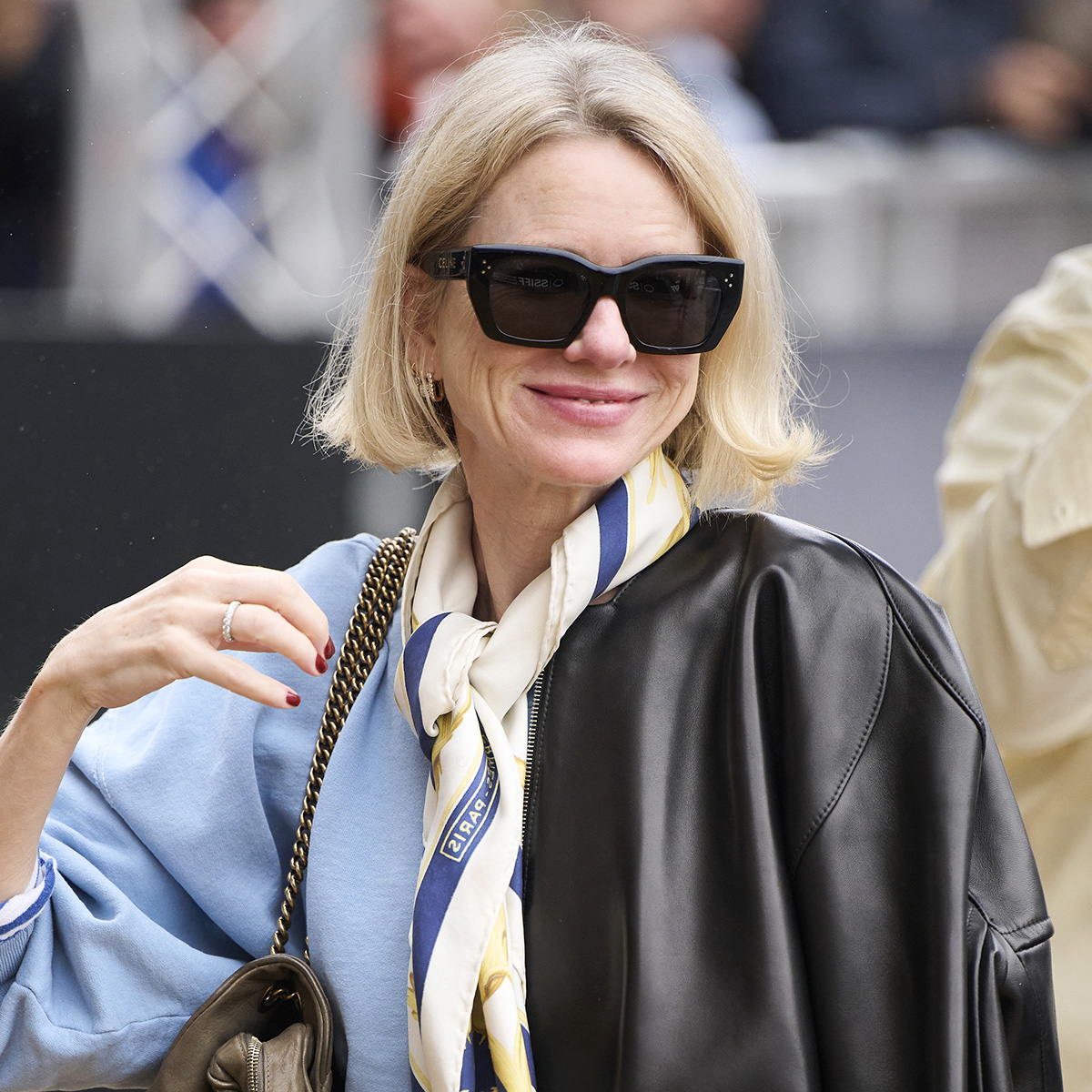 Naomi Watts in in San Sebastian, Spain wearing a leather bomber jacket, neck scarf, light blue sweater, bootcut jeans, and Adidas Samba sneakers