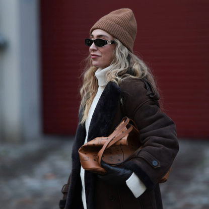 BERLIN, GERMANY - NOVEMBER 24: Sonia Lyson seen wearing a long dark brown shearling Ducie coat layered over wide, flowing cream trousers from Review and COS. The look is completed with soft slip-on shoes from Zara and a ribbed beige COS beanie. Accessories include a structured tan MCM tote bag with monogram patterning and slim black Gucci sunglasses. The outfit blends cozy winter textures with elevated neutral tones on November 24, 2025 in Berlin, Germany. (Photo by Jeremy Moeller/Getty Images)