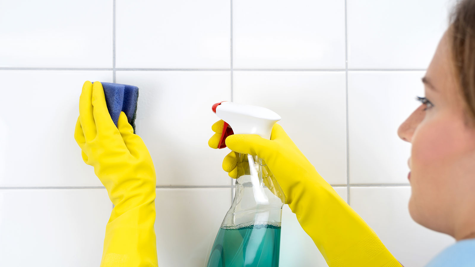 Woman in yellow rubber gloves spraying solution onto white tiles and grout and cleaning with a blue kitchen scrubber