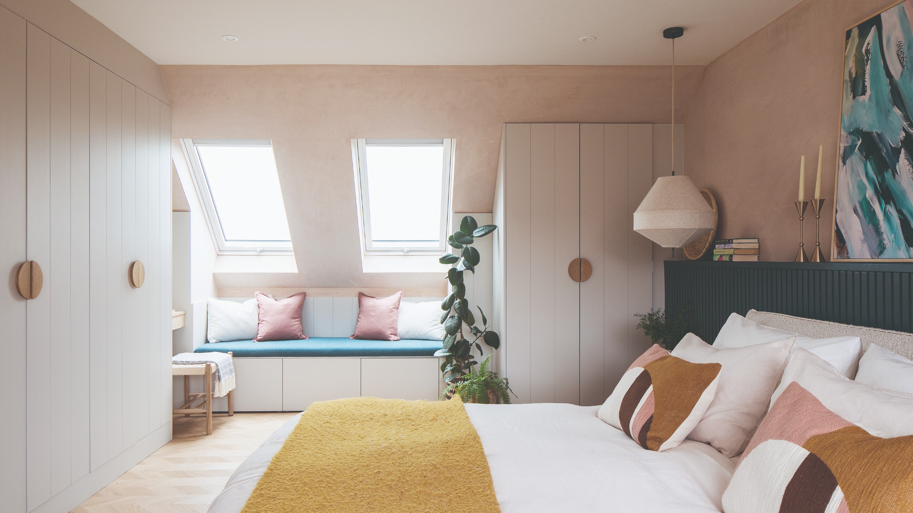 A plaster pink-painted bedroom with a slanted ceiling, a window seat and built-in cream wardrobes