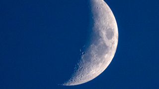 The moon is pictured against a dark blue sky with a thin crescent on its right side lit by direct sunlight.
