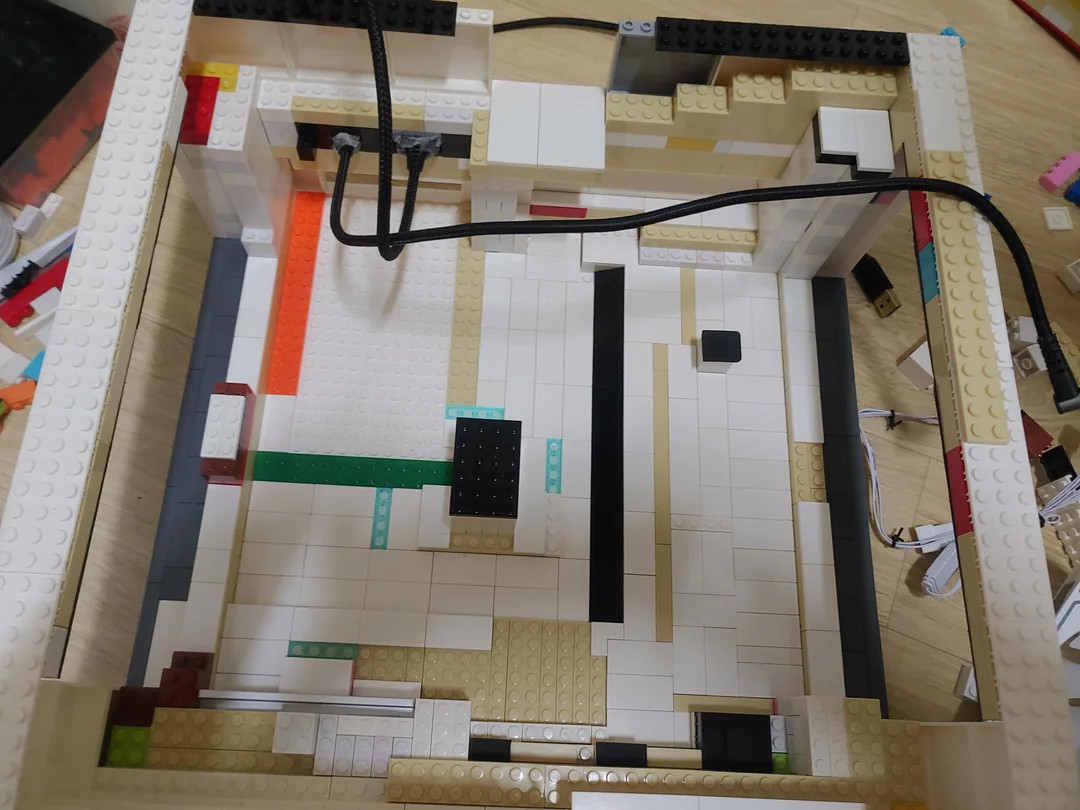 LEGO PC case with white, black, orange, and green bricks forming a rectangular shape on a wooden surface. Black wires cross the setup.