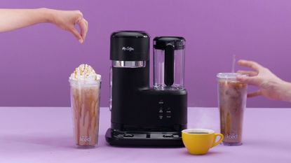 Mr. Coffee single-serve frappe, iced, and hot coffee maker on purple background. A hand on the left is sprinkling chocolate on a creamy drink and the one on the right is grabbing an iced latte with a straw