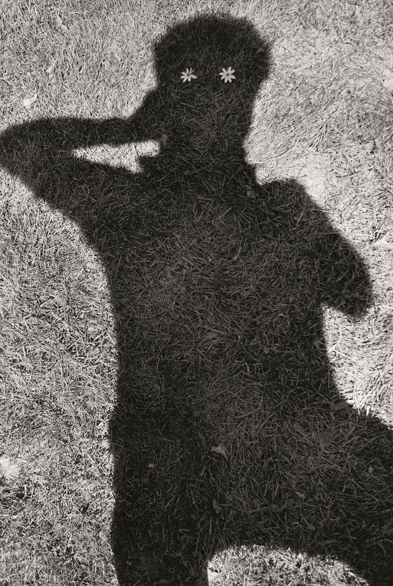 Black. andwhite image of a shadow of a person making a peace sign on grass, adorned with two small daisies where their eyes would be