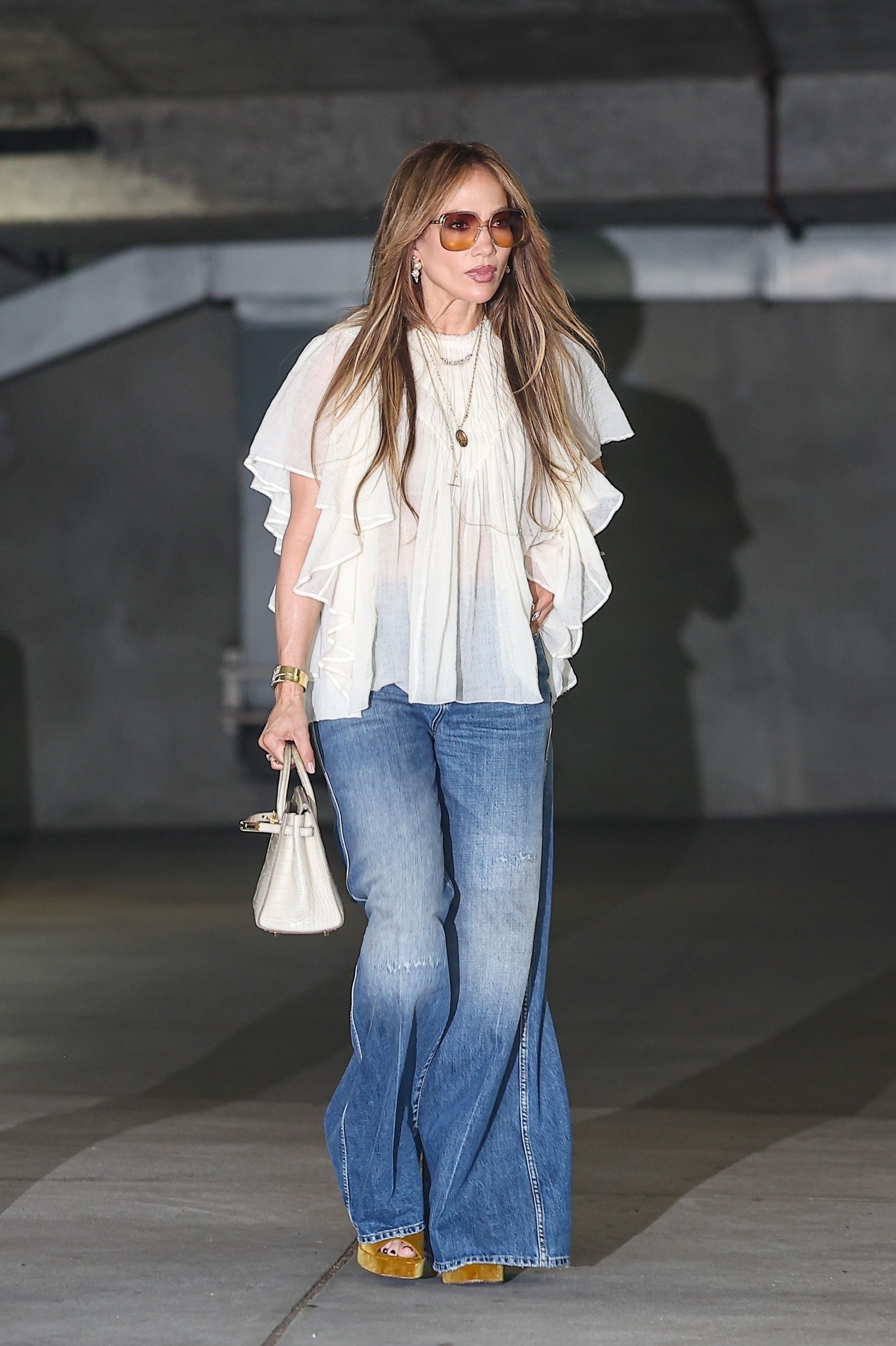 JLo wears jeans with a boho blouse and velvet platform heels. She carries a white bag in her hand and walks through a car park.