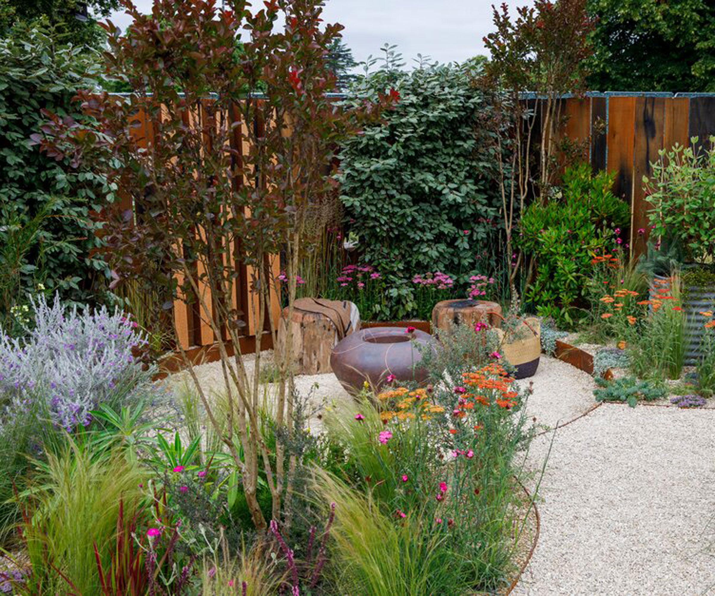 small garden design with deep flowerbed including plants, flowers, ornamental grasses and a multi-stem tree, fire pit and seats, fence and gravel landscaping