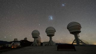 long view of three mushroom-shaped telescopes underneath galaxies in the night sky