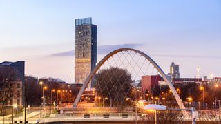 A night time shot of the Manchester skyline, with a highrise building in the backgroud and the arc of the hulme bridge in the foreground