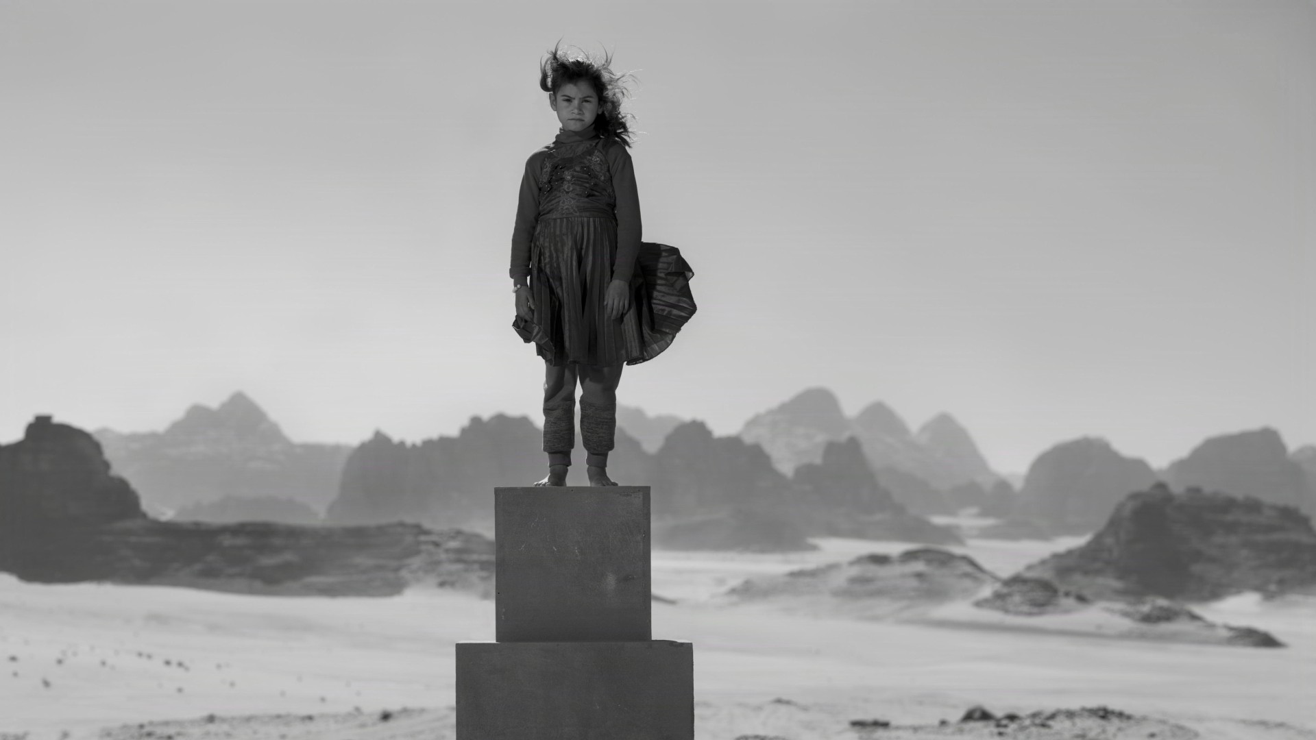 A young girl stands defiantly atop a tall pedestal of stacked blocks in the middle of a barren desert.