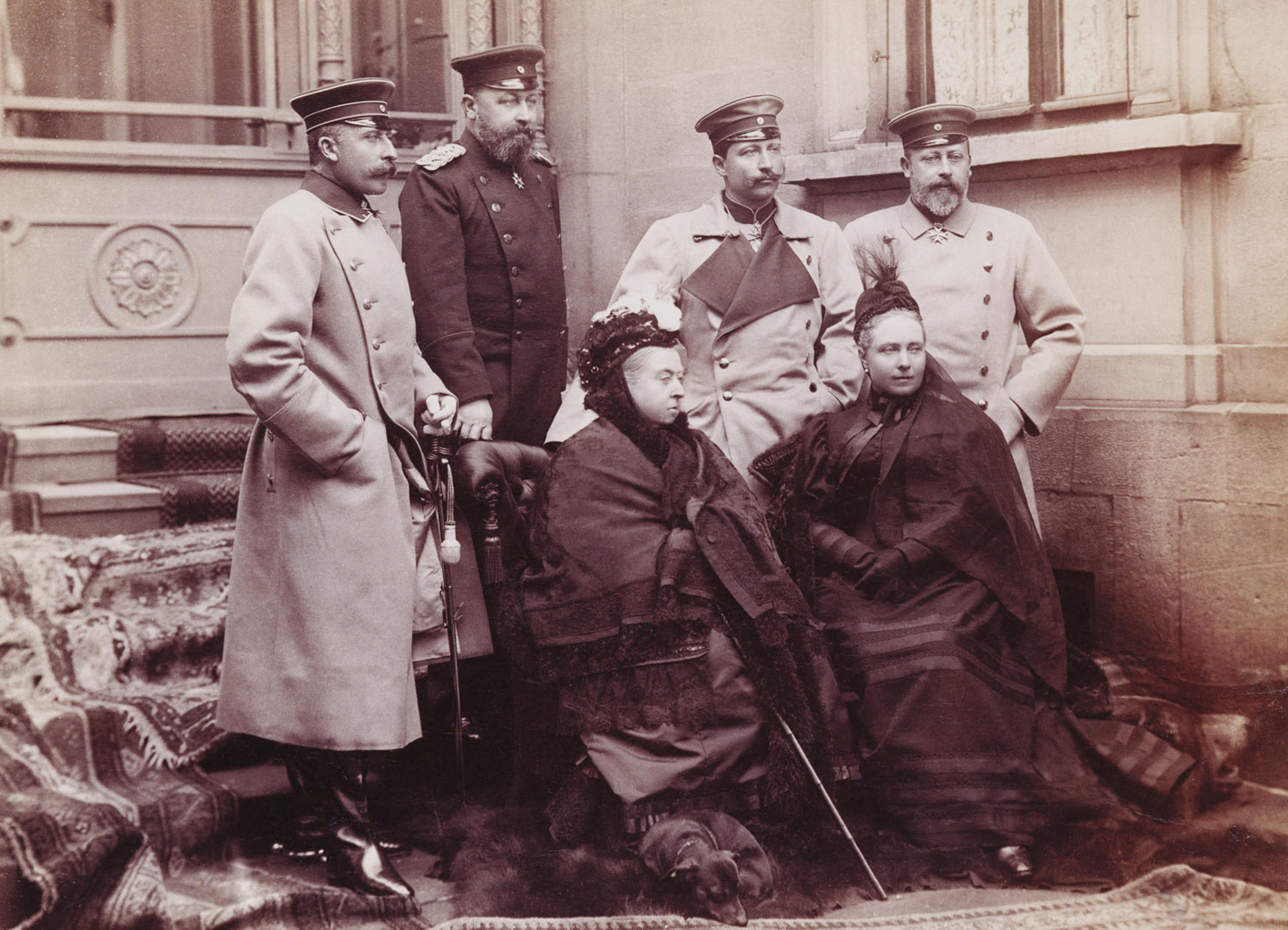 A sepia-toned 1894 group photograph of European royalty gathered outside a grand building in Coburg, with Queen Victoria seated at the centre and a dachshund lying at her feet, surrounded by formally dressed royal relatives in military coats and Victorian dress.