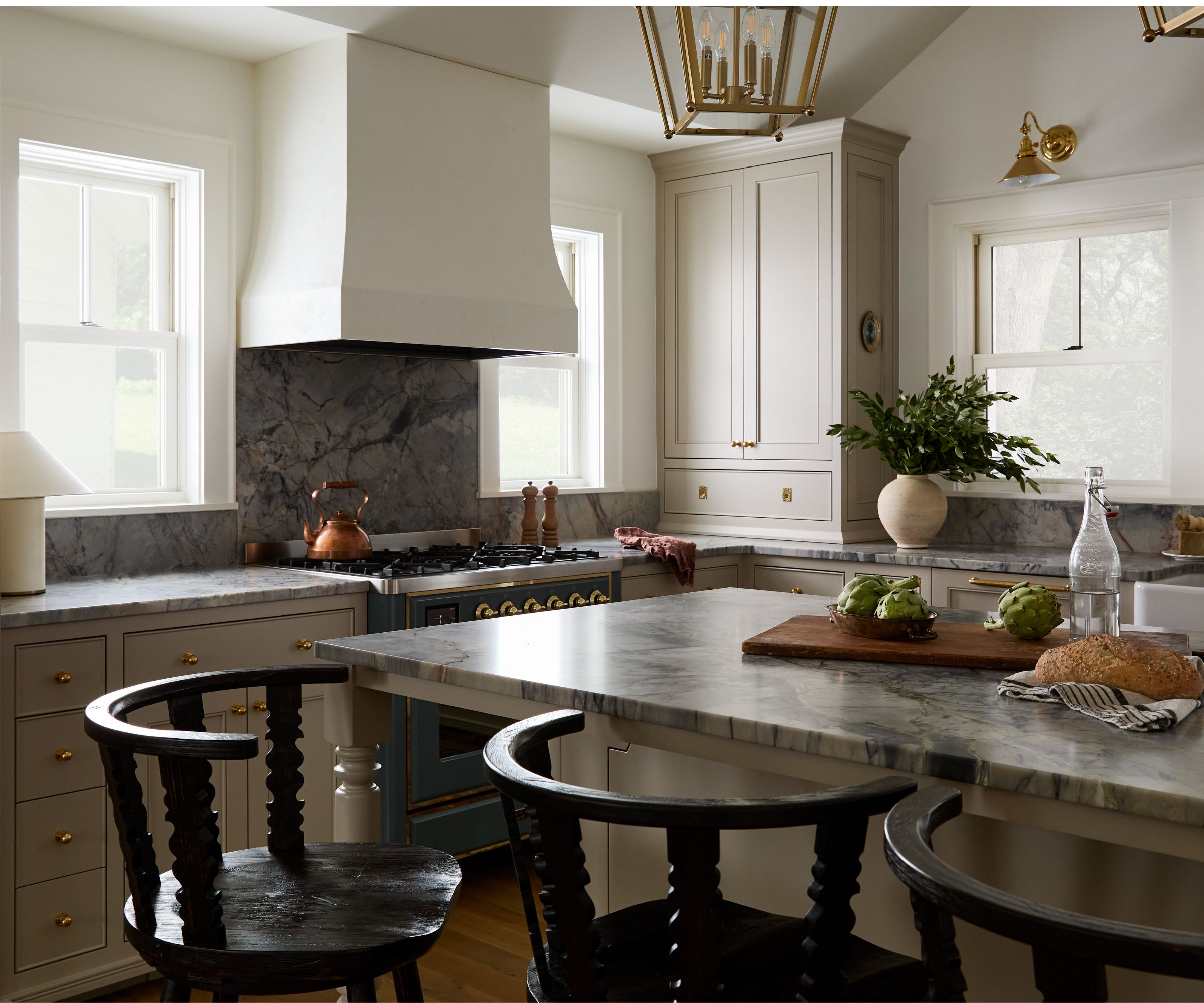 earthy neutral kitchen with large windows out to the lakeside view, a large plaster cooker hood, and blue marble countertops