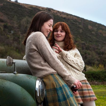 Hazel Doupe as Marian Price and Lola Petticrew as Dolours Price, leaning against the front of an old green car covered in dirt, in 'Say Nothing.'