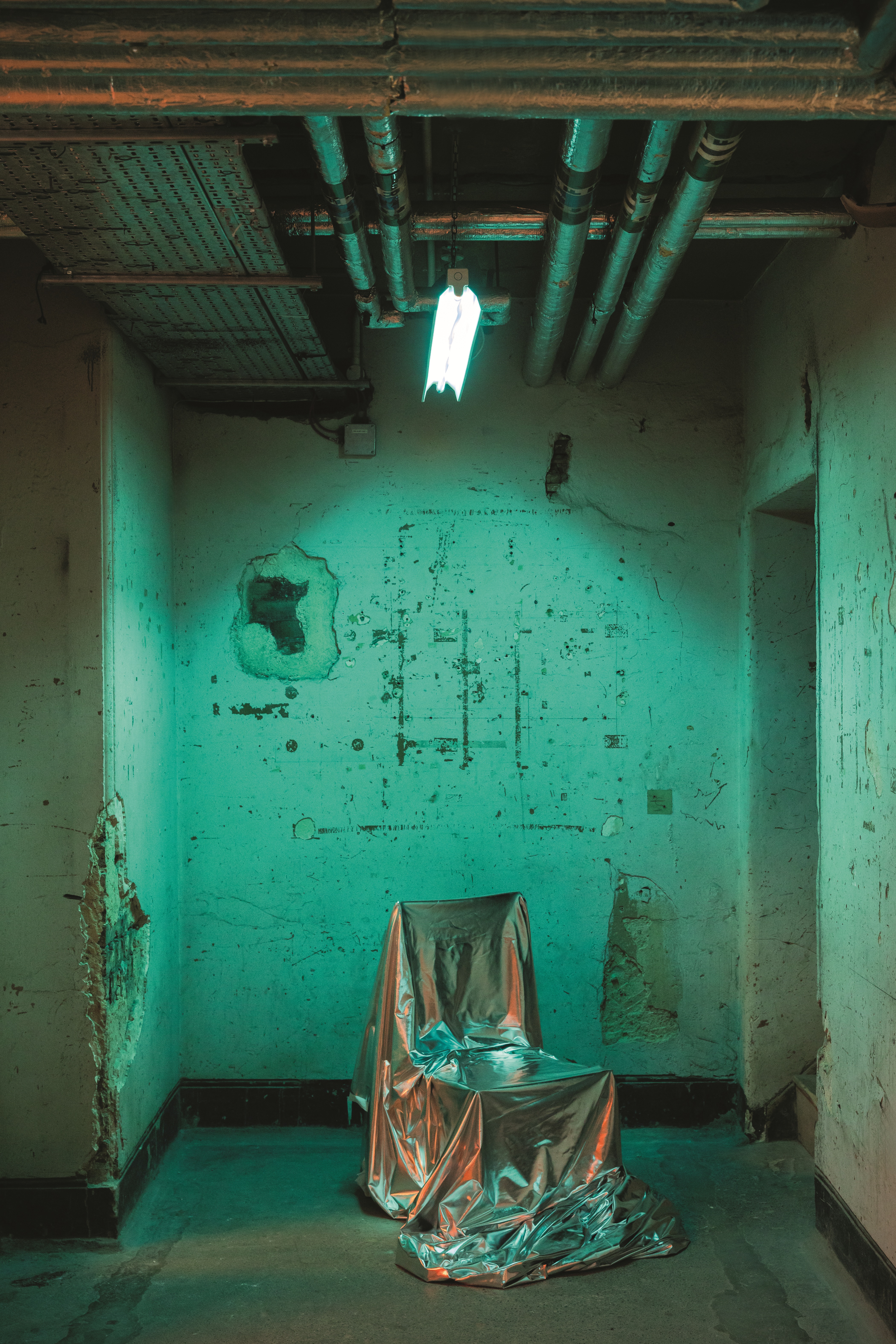 A neon-lit, water green-hued industrial room with unfinished walls and chrome ventilation pipes houses a chair covered in a silvery fabric.