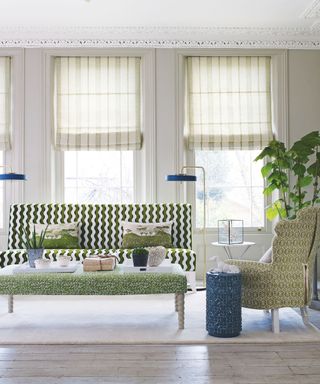 A bright living room with large windows with geometric blinds, and patterned geometric sofa, armchair and padded footrest in green patterns, two free-standing gold and blue lamps, a blue cylindrical coffee table, and a large plant