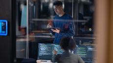 Female software engineer working on a desktop computer in an office space while male colleague stands over desk checking work on a tablet.