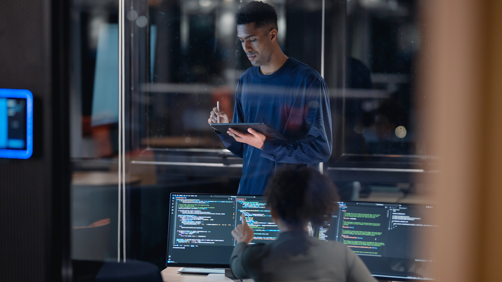 Female software engineer working on a desktop computer in an office space while male colleague stands over desk checking work on a tablet.