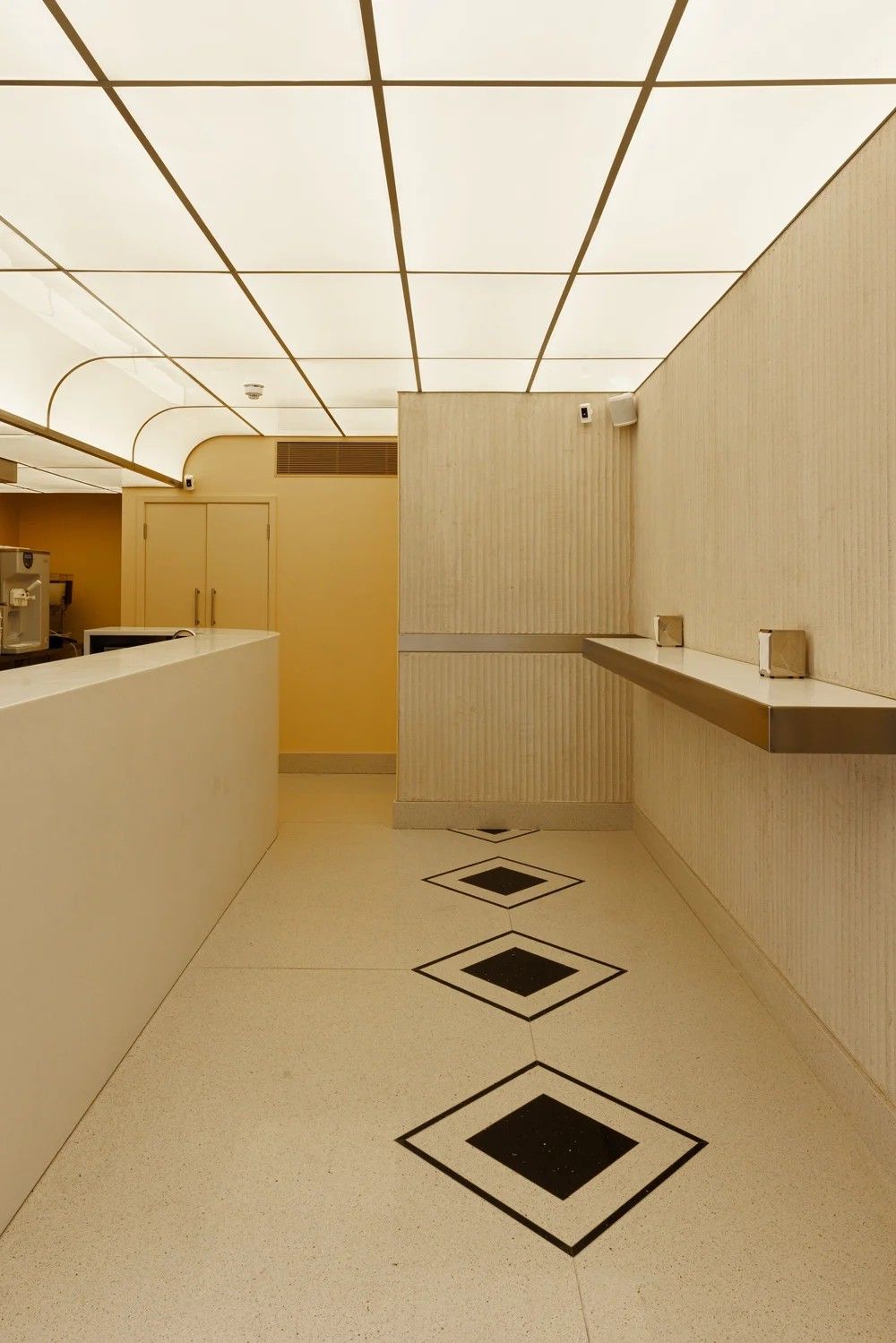 A retro-fueled diner features coffered, back-lit ceilings with brass detailing, terrazzo floors and walls in buttery tones, with textural accents, a wood and a pale yellow wall.