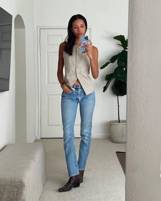 influencer and model Tylynn Nguyen poses for a mirror selfie wearing a tan linen vest, low-rise skinny jeans, and Western boots