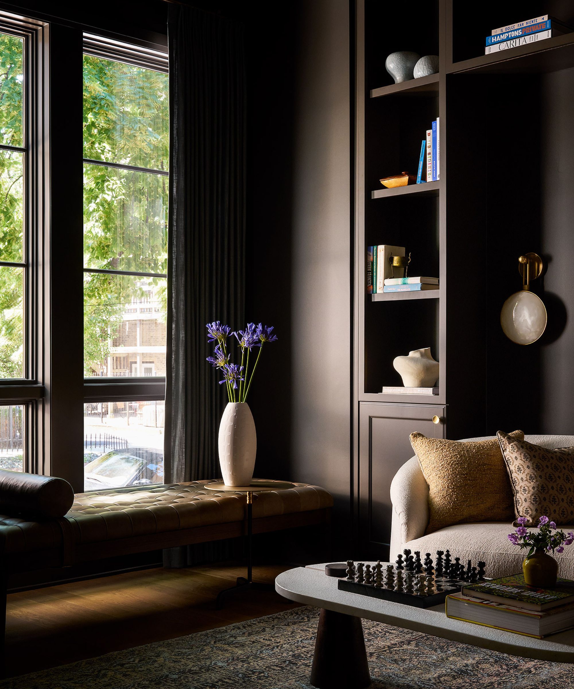 A study with dark brown color-drenched walls and joinery, with a floor-to-ceiling window, a leather window bench, a beige modern armchair with caramel-toned cushions, and a modern rounded coffee table.