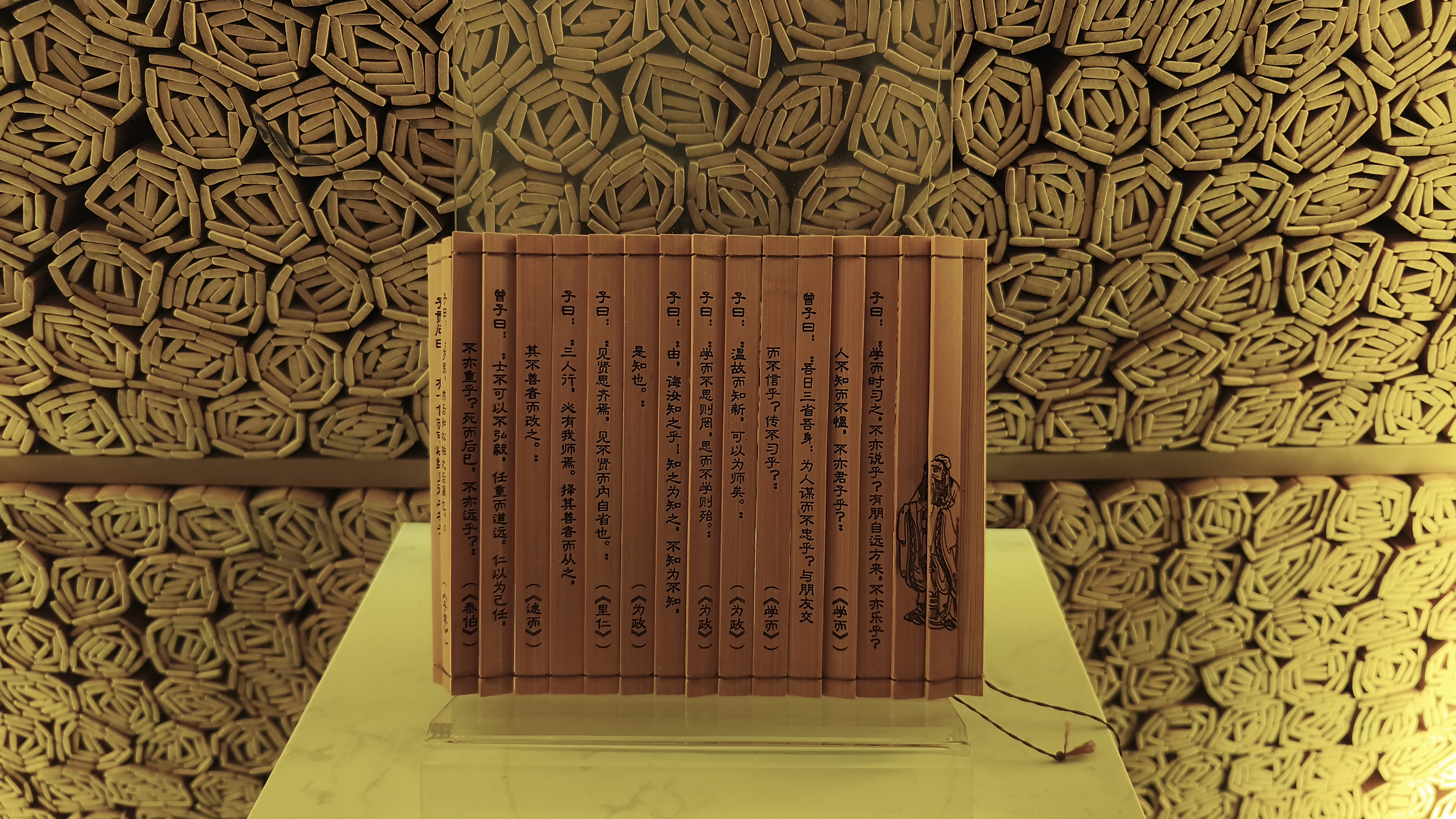An accordion-style wooden book featuring traditional Chinese calligraphy is displayed on a clear stand in front of a wall constructed from thousands of rolled bamboo scrolls.