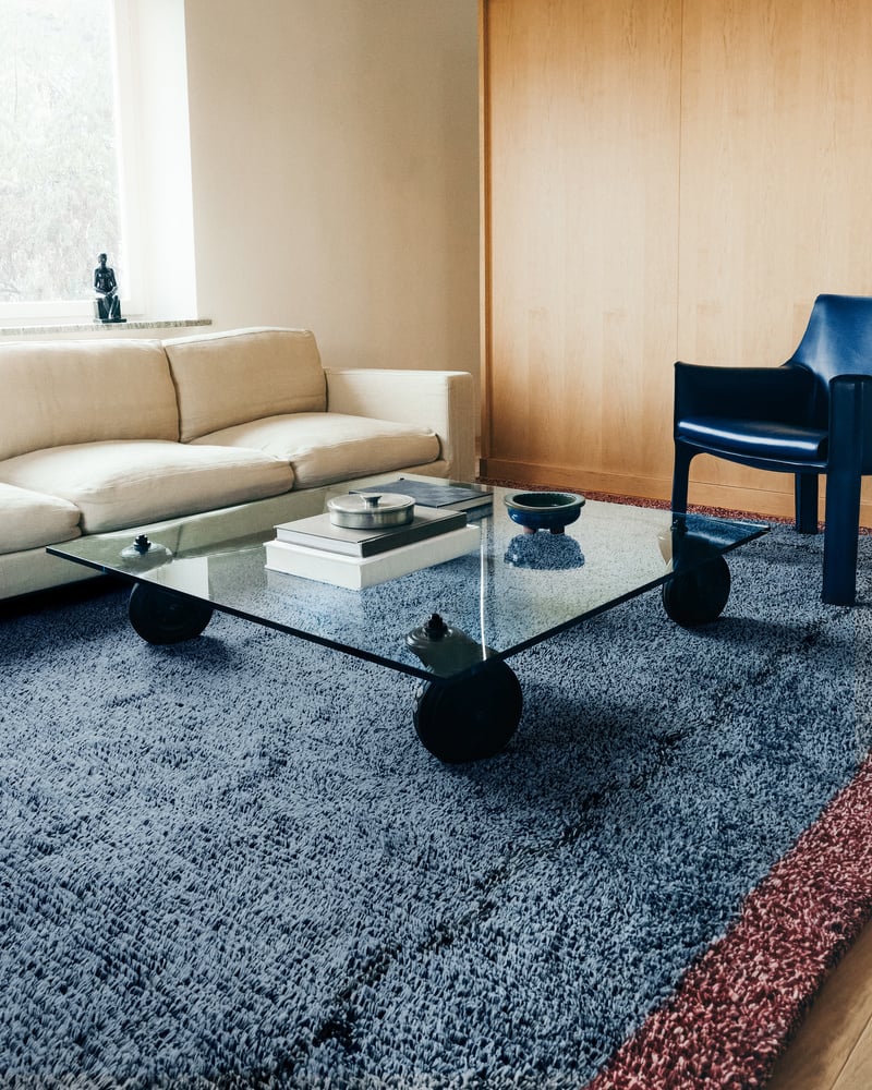 Image of a modern, Scandinavian-inspired living room with a white sofa and a blue short pile shag rug with a red border. There is a square, glass coffee table in the center of the rug and a blue leather accent chair.
