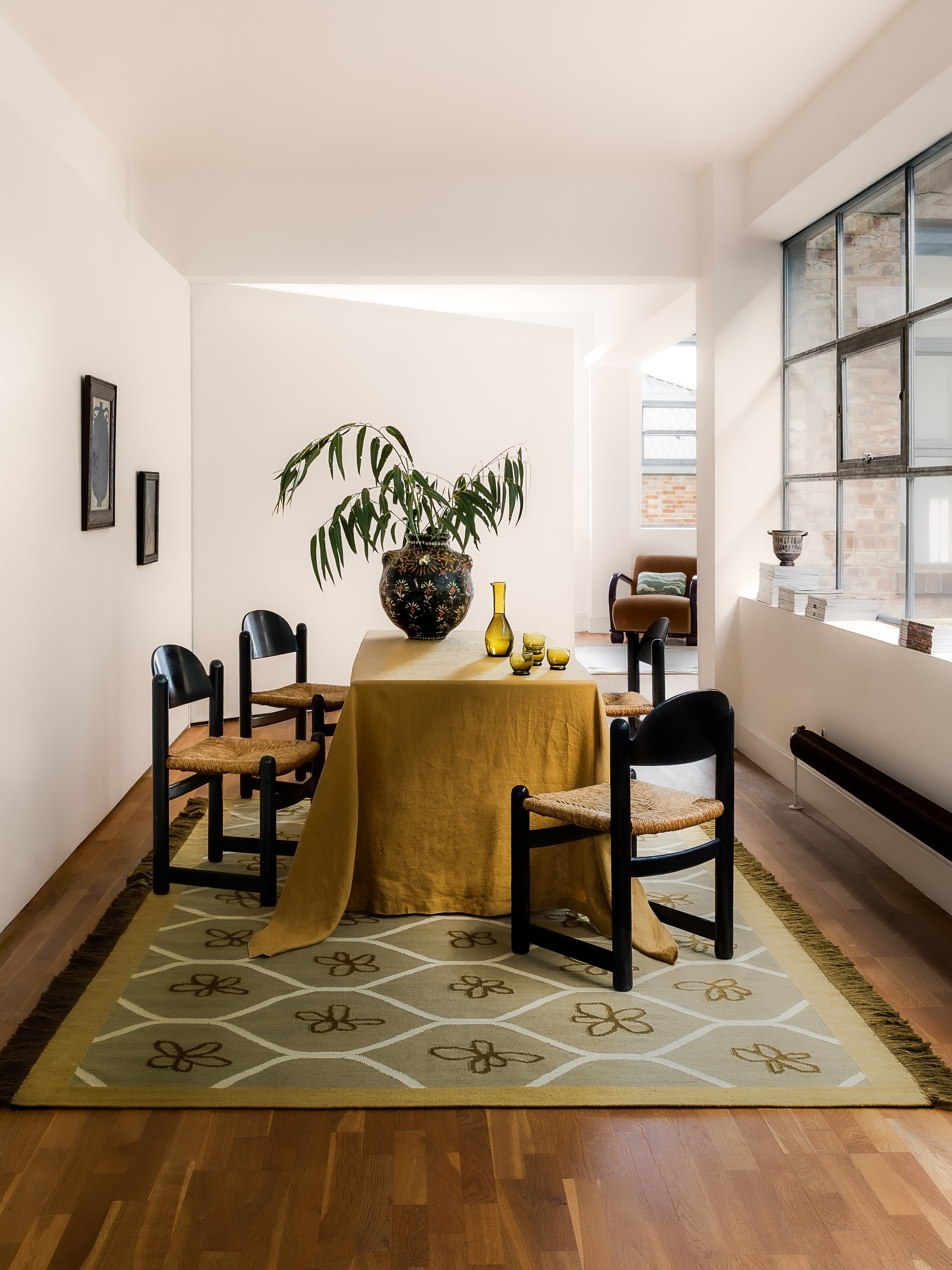 Image of a white dining room with wooden floors and a green patterned rug on the floor. There's a dining table with a mustard table cloth and black and rattan dining chairs.