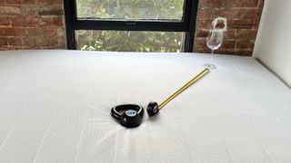Photo of Helix Midnight mattress in a bedroom, with a weight on it, and a wine glass stood upright on the mattress surface nearby. A tape measure shows the distance between glass and weight.