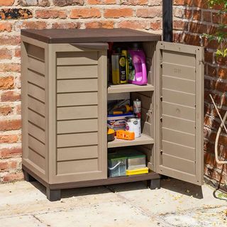 Brown plastic storage cabinet with 2 shelves standing on paving next to red brick wall