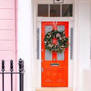 Festive wreath hanging from ribbon on a red wooden front door of a pink building