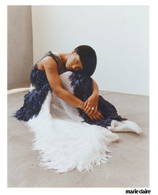 Actress Myha'la Herrold sitting on a concrete floor with her knees to her chest in a blue and white feather dress