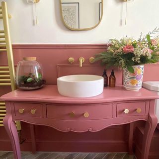 Upcycled vanity table in gorgeous bathroom scheme, finished in rose paint, with round white sink, brass hardware, terrarium, and florals