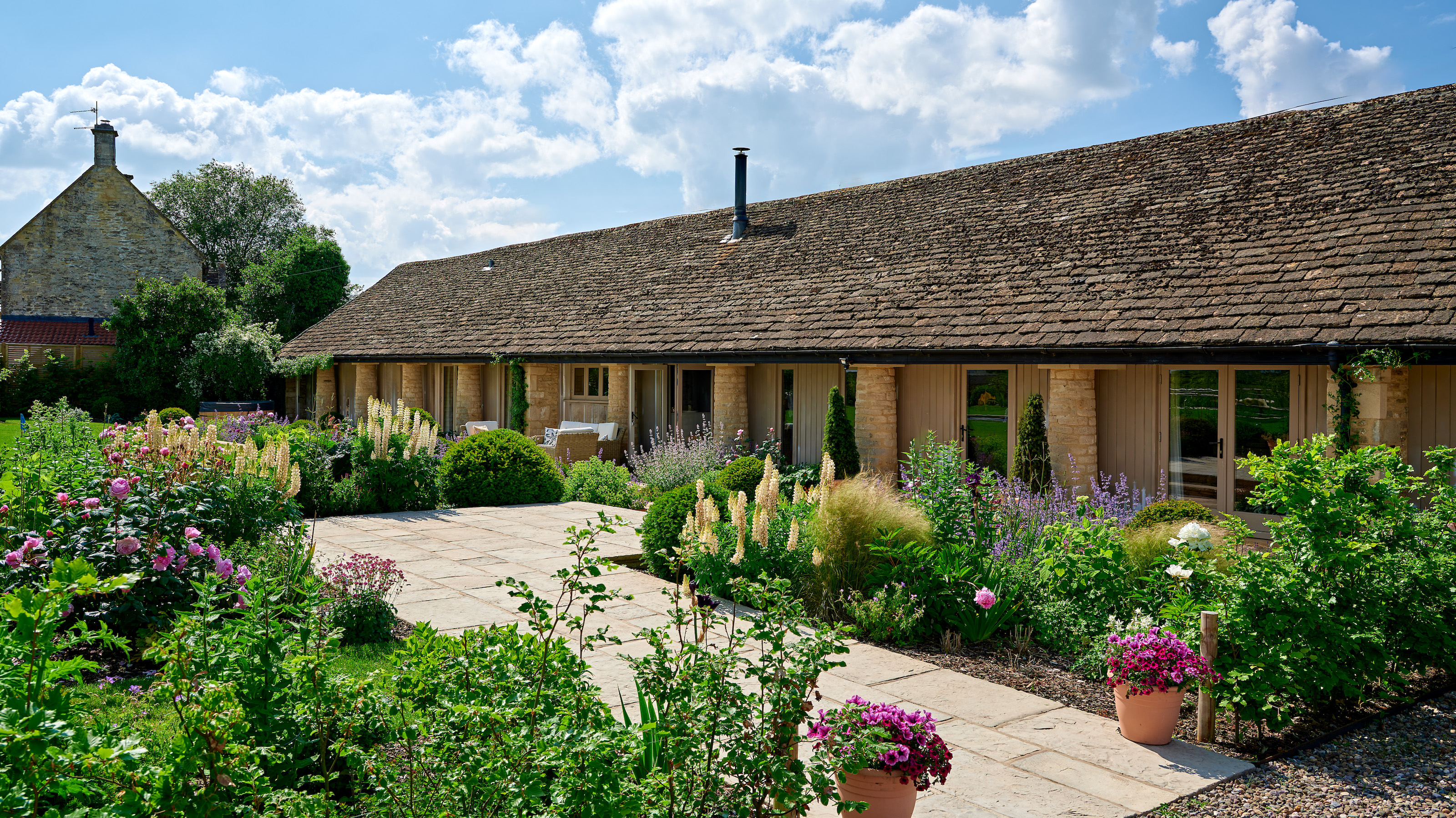 exterior of single storey converted cowshed with paving surrounded by country cottage flowers