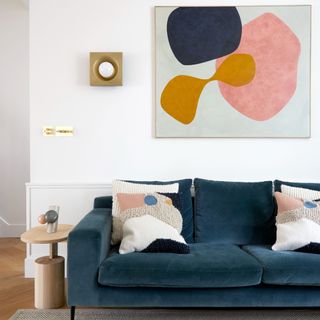 A living room with a blue velvet sofa with an artwork hanging above it and a wall sconce next to it