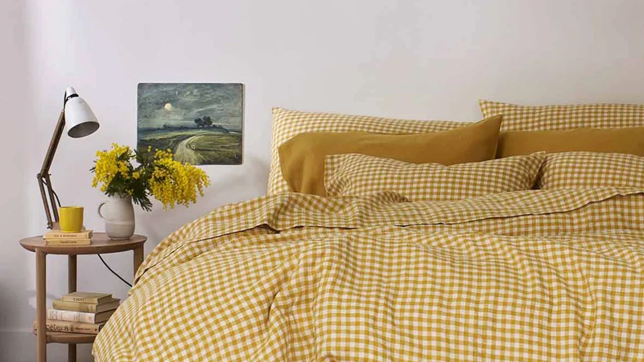 A bedroom with a large bed covered in a yellow gingham duvet and matching pillowcases. A wooden bedside table holds a lamp and a vase with yellow flowers