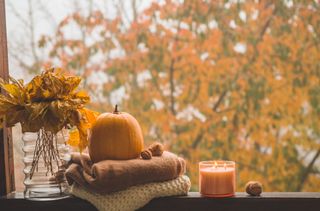 A window with a pumpkin, an orange candle, a conker, some autumn foliage, and some brown and cream jumpers rolled up.