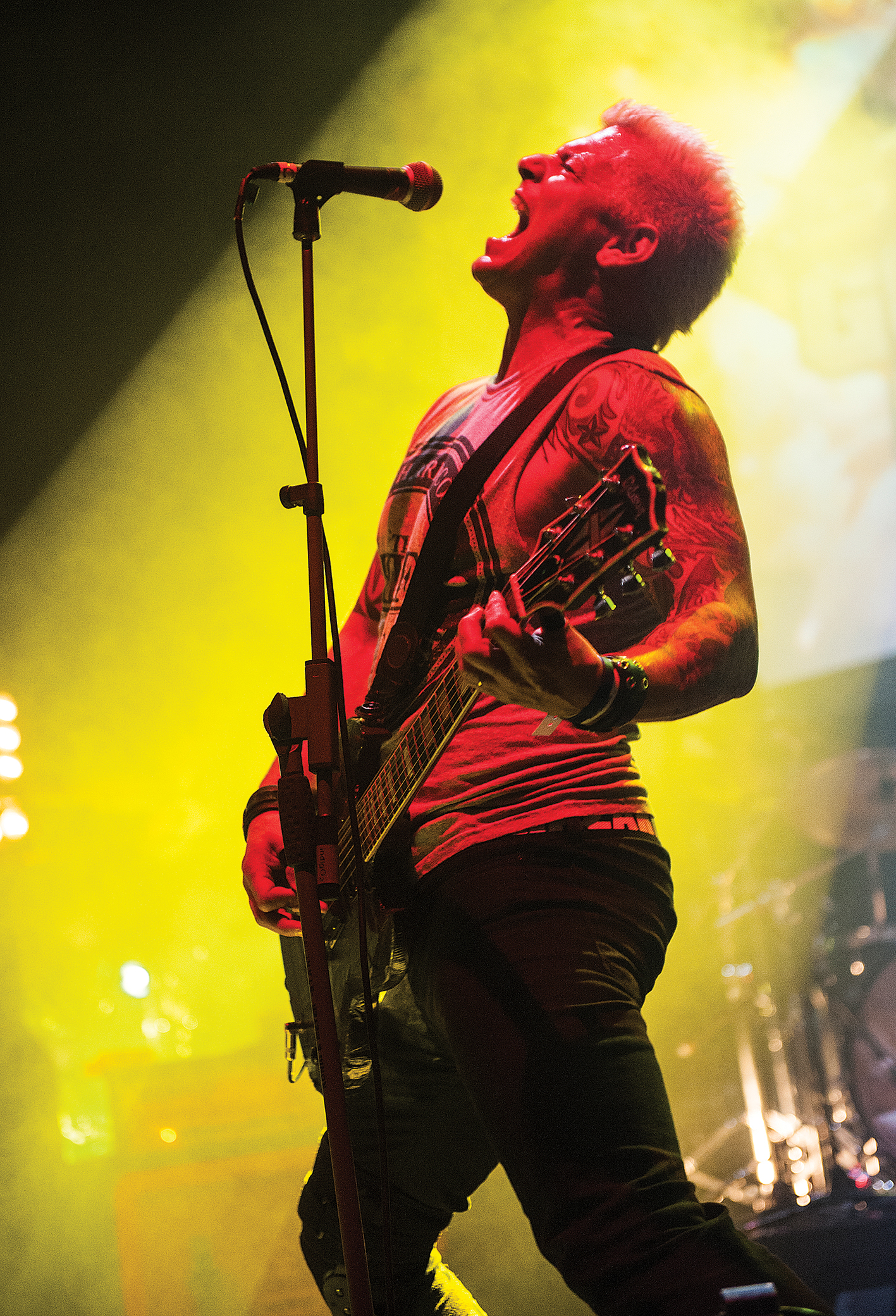 A guitarist passionately performs on stage, illuminated by vibrant yellow lighting, showcasing intricate tattoos and an electric guitar